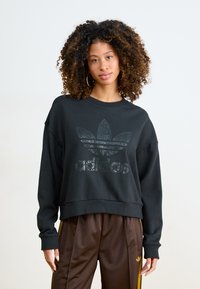 Fekete, rövidített pamut pulóver, túlméretezett ujjakkal, elöl texturált Adidas logóval. Barna selyem nadrággal párosítva.