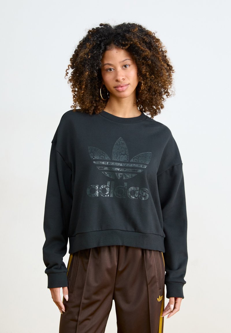 Fekete, rövidített pamut pulóver, túlméretezett ujjakkal, elöl texturált Adidas logóval. Barna selyem nadrággal párosítva.