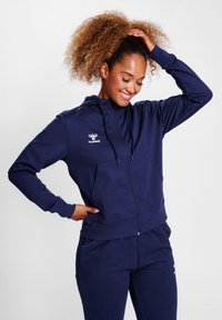 Marineblauwe zip-up hoodie van zacht materiaal, met een voorvak, elastische manchetten en logo detail op de linkerkant van de borst. Gecombineerd met bijpassende broek.
