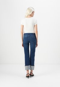 Denim jeans in dark blue with a light denim cuff. Features a straight cut, back pockets, and a simple waistband. Paired with a white top.