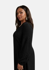 Černé šaty s dlouhým rukávem, vyšívanými vzorci spirál na rukávech, vyrobené z měkké tkaniny; mají přiléhavé živůtek a splývavou siluetu.