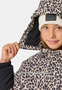 Leopardenmuster-Jacke in Beige mit schwarzen Akzenten, ausgestattet mit einem Reißverschluss an der Kapuze, Druckknopfverschluss und elastischen Bündchen. Darunter eine beige Strickmütze.