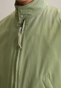 Lichtgroene jas met een hoge kraag, twee drukknoopjes en een ritssluiting aan de voorkant. Heeft een stoffen strik om de hals en zijzakken.