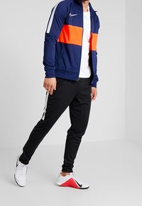 Veste zippée bleu marine avec une bande horizontale orange, accents blancs ; associée à un pantalon noir avec des rayures blanches sur les côtés et des chaussures de sport blanches et rouges.