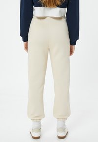 Cremefarbene Jogginghose mit schmal zulaufendem Schnitt, elastischem Bund und Knöchelbündchen, aus weichem Stoff, kombiniert mit weißen Sneakers und Socken.