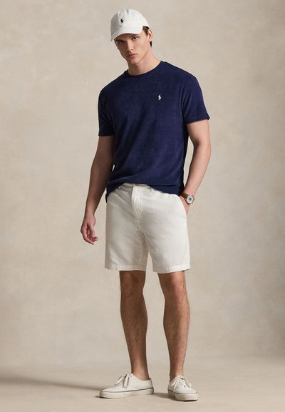 Μπλε ναυτικό κοντομάνικο μπλουζάκι με μικρό λογότυπο, λευκό βαμβακερό σορτς, λευκό καπέλο και λευκά αθλητικά παπούτσια. Απλός σχεδιασμός, άνετη εφαρμογή και ελαφρύ ύφασμα.