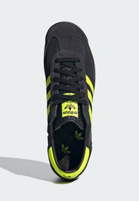 Černá semišová sportovní bota se třemi výraznými neonově žlutými pruhy, texturovanými černými tkaničkami a polstrovaným límcem. Výrazné logo na jazyku.