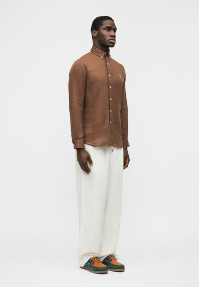 Άνδρας στέκεται με το πρόσωπο προς τα εμπρός, φορώντας ένα καφέ πουκάμισο με κουμπιά, φαρδιά λευκά παντελόνια και πολύχρωμα παπούτσια με κορδόνια, μπροστά σε απλό φόντο.