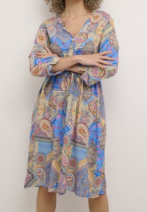 Kobieta ubrana w sukienkę z długim rękawem sięgającą kolan, w niebieski wzór paisley i geometryczny, z rękami skrzyżowanymi na wysokości talii.