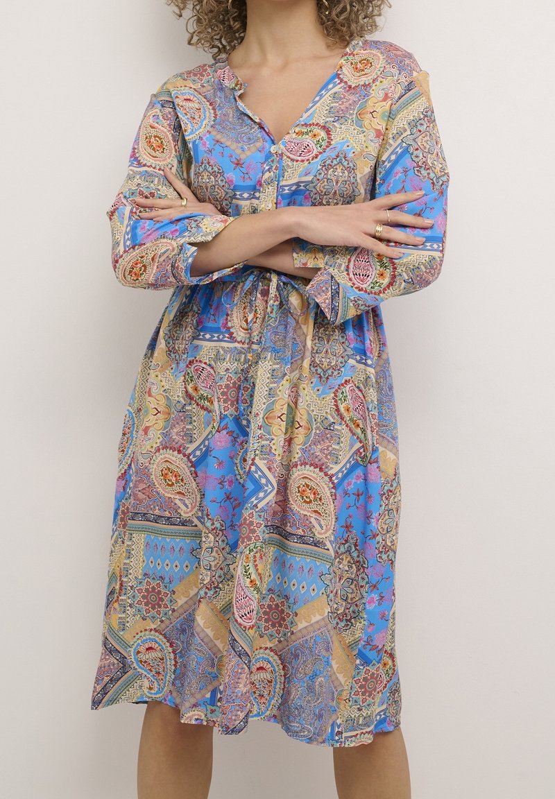 Femme portant une robe longue à manches longues, arrivant au genou, avec un motif paisley bleu et géométrique, les bras croisés au niveau de la taille.