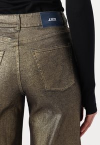 Vaqueros de mezclilla dorados metálicos con un diseño de pierna recta. Presenta dos bolsillos traseros y una etiqueta de marca azul en la cinturilla. Textura suave.