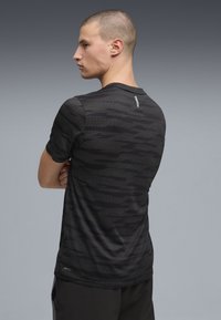 Νεαρός με ξυρισμένο κεφάλι που φοράει μαύρο αθλητικό πουκάμισο με μοτίβο, φαίνεται από πίσω με τα χέρια σταυρωμένα, στέκεται μπροστά σε γκρι φόντο.