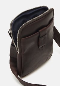 Braune Ledertasche mit Reißverschluss, marineblauem Innenraum, Vordertasche und gewebtem Schultergurt. Mit einem dekorativen Verschlussdetail.
