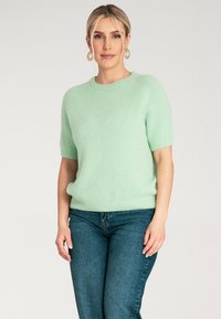 Jasnozielony sweter z krótkim rękawem, wykonany z miękkiego, puszystego materiału, z okrągłym dekoltem i luźnym krojem, w zestawieniu z ciemnoniebieskimi dżinsami.