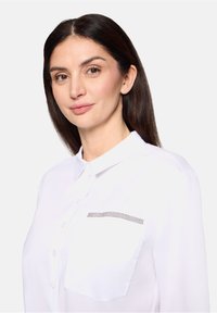Chemise blanche à boutons avec une poche ornée d'une rangée de décorations. Tissu lisse, col classique et manches longues.