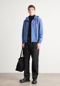 Blå hettegenser med glidelås og logo-patch, svart høyhalset genser, svarte jeans med synlig søm, og svarte slip-on sko, holder en svart totebag.