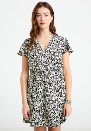 Kobieta o długich brązowych włosach, ubrana w krótką czarno-białą sukienkę w kwiaty i srebrną bransoletkę, stojąca na tle jednolitego białego tła.