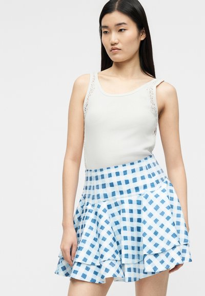 Γυναίκα που φοράει λευκό αμάνικο τοπ με δαντέλα στα πλάγια και φούστα με βολάν σε μπλε και λευκό καρό, στέκεται μπροστά σε ένα απλό φόντο.
