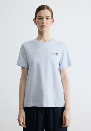 Mladá žena s vlasy staženými dozadu má na sobě světle modré tričko značky GANT a tmavé kalhoty, stojí před jednobarevným bílým pozadím.