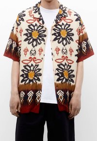 Chemise à manches courtes avec une base crème, ornée de motifs floraux multicolores et abstraits en rouge, orange et noir. Fermeture à boutons sur le devant.