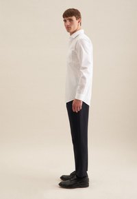 Junger Mann steht seitlich, trägt ein weißes Langarmhemd, schwarze Hose und schwarze Lederschuhe vor einem einfarbigen Hintergrund.