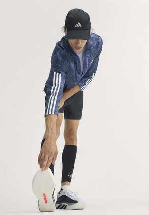 Personne en tenue de sport se penche en avant pour toucher la semelle de sa chaussure blanche, portant une casquette noire, une veste bleue avec des bandes blanches et un short noir.