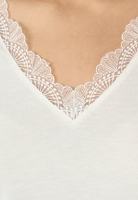 Witte V-halsblouse met een delicate kantafwerking bij de hals. De stof is glad met een subtiele textuur, die de kantdetails accentueert.