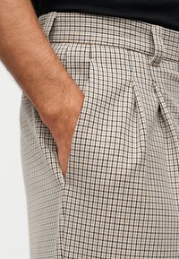 Χέρι στην τσέπη μπεζ παντελονιού με μαύρο και καφέ καρό σχέδιο, εμφανίζοντας πιέτες και λεπτομέρεια θηλιάς για ζώνη.