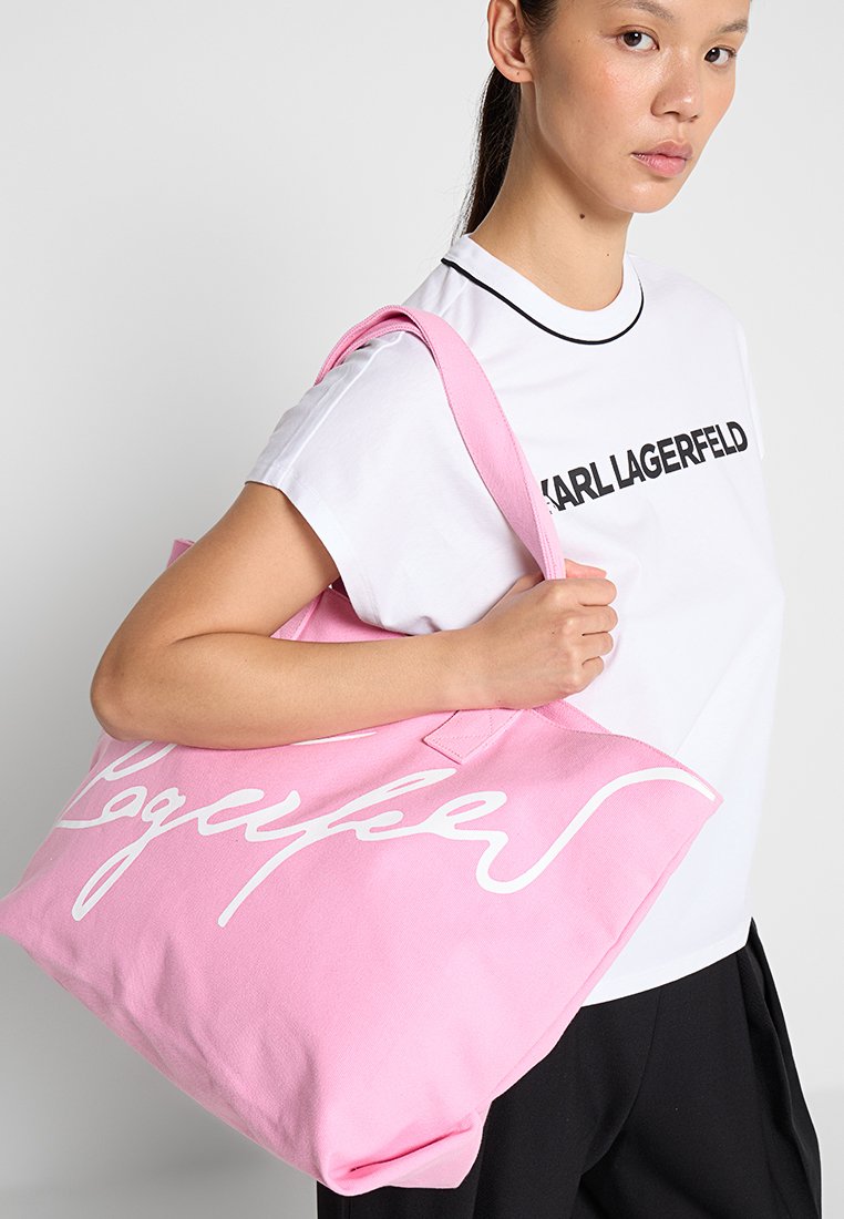 Jeune personne tenant un grand sac fourre-tout rose avec un script blanc, portant un t-shirt blanc Karl Lagerfeld et un pantalon noir sur un fond uni.