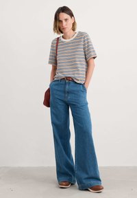 Camiseta a rayas con cuello redondo, que presenta múltiples líneas horizontales en marrón, naranja y azul. Vaqueros de pierna ancha azules, zapatos marrones. Bolso rojo.