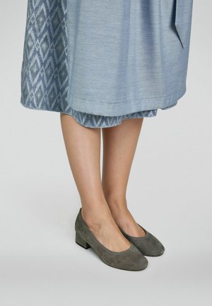 Jambes portant des chaussures grises en daim à petit talon, posées sur une surface unie, avec une jupe bleue mi-longue dotée d'une couche intérieure à motifs.