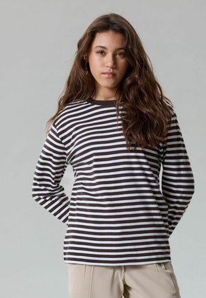 Jonge vrouw met lang bruin haar, die een zwart-wit gestreept shirt met lange mouwen en beige broek draagt, staat tegen een lichtgrijze achtergrond.