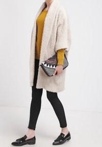 Beige, pluizige, oversized jas; gele lange mouwen shirt; zwarte skinny broek; zwarte instappers; geprinte clutch met geometrische ontwerpen.