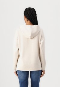Hoodie bege claro com uma textura suave, corte solto e um ventilador nas costas. Combinado com jeans de sarja azul. Apresenta um capuz com cordão.