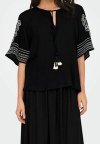 Femme portant un blouse noire amples avec des motifs brodés blancs sur les manches et des liens à franges au col, associée à une jupe noire.
