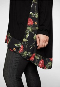 Zwarte top met bloempatroon langs de zoom, met rode rozen en groene bladeren op een donkere achtergrond. Kenmerkt zich door een losse pasvorm.