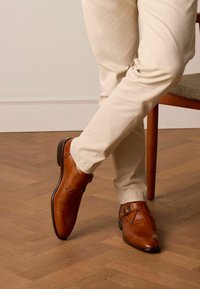 Braune Lederschuhe mit einem eleganten Design, versehen mit einem seitlichen Schnallenetail, getragen mit hellen Hosen, auf einem Holzboden.