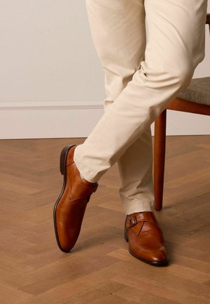 Des chaussures en cuir marron au design épuré, avec un détail de boucle sur le côté, portées avec un pantalon de couleur claire, sur un sol en bois.