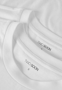 Dve beli bombažni majici, zloženi ena na drugo, z okroglimi izrezki in etiketi, na katerih je z napisom "TWO SOON" in "S" v preprosti črni pisavi.