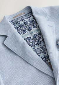 Lichtblauwe blazer met textuur en gedetailleerde voering met bloemenpatroon in marineblauw, wit en beige, zichtbaar aan de binnenkant van de kraag.