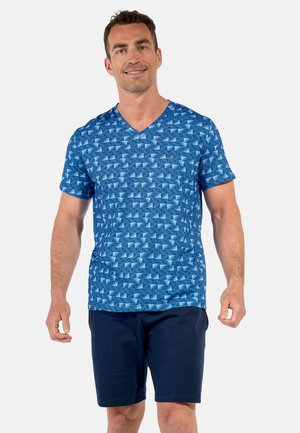 Mann trägt blau gemustertes kurzärmeliges V-Neck-Shirt und einfarbige dunkelblaue Shorts, steht vor einem schlichten hellen Hintergrund.