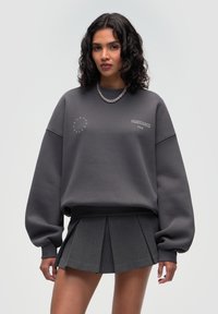 Grauer Sweatshirt mit übergroßen Ärmeln, das ein dezentes Logo-Detail aufweist. Kombiniert mit einem kurzen, plissierten grauen Rock. Strukturierter Stoff.