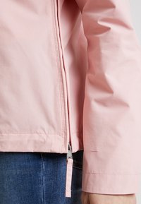 Jasnoróżowa kurtka o gładkiej, błyszczącej fakturze, z zapięciem na zamek i dopasowanymi mankietami, zestawiona z niebieskimi dżinsami.