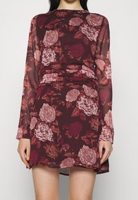 Långärmad klänning i bordeauxfärgad tyg med blommönster i nyanser av rosa och mörkröd, med rynkad midja och lager-på-lager textur.