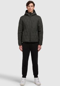 Dark green quilted jacket with a hood, featuring a geometric pattern, zippered chest pocket, and elastic cuffs. Paired with black pants and white sneakers.