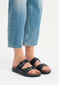 Czarne skórzane sandały z dwoma szerokimi paskami i złotymi klamrami, posiadające wyściełaną wkładkę i teksturowaną czarną podeszwę, noszone z przykrótkimi niebieskimi dżinsami.