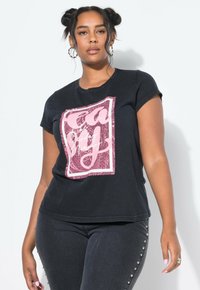 Mujer con dos moños, vistiendo una camiseta negra con texto gráfico rosa y pantalones negros con tachuelas plateadas a los lados, contra un fondo blanco.
