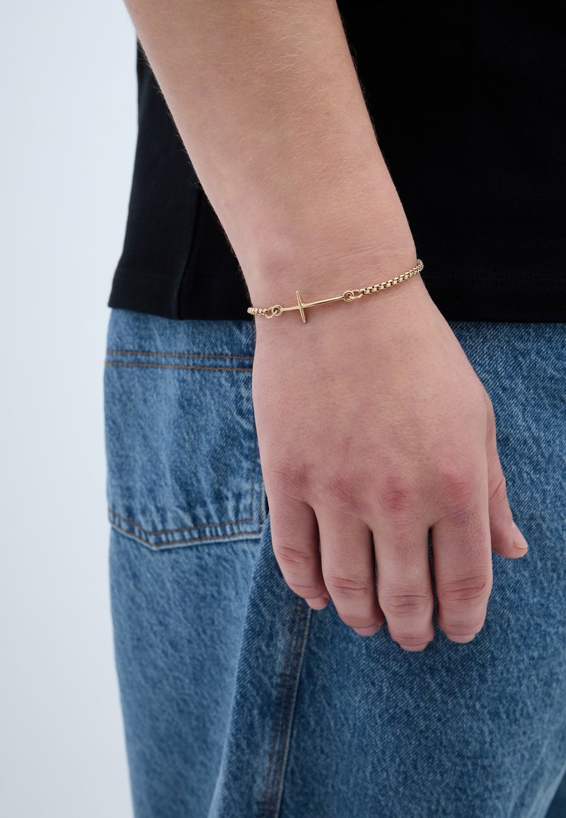 Arany lánc karkötő vízszintes sáv díszítéssel, amely egy csuklón van viselve. A kéz egy kék farmeranyaghoz ér. Sima textúra.