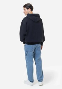 Sweatshirt preto com capuz, corte descontraído, combinado com jeans largos azuis claros com bolsos laterais, usado com ténis brancos.