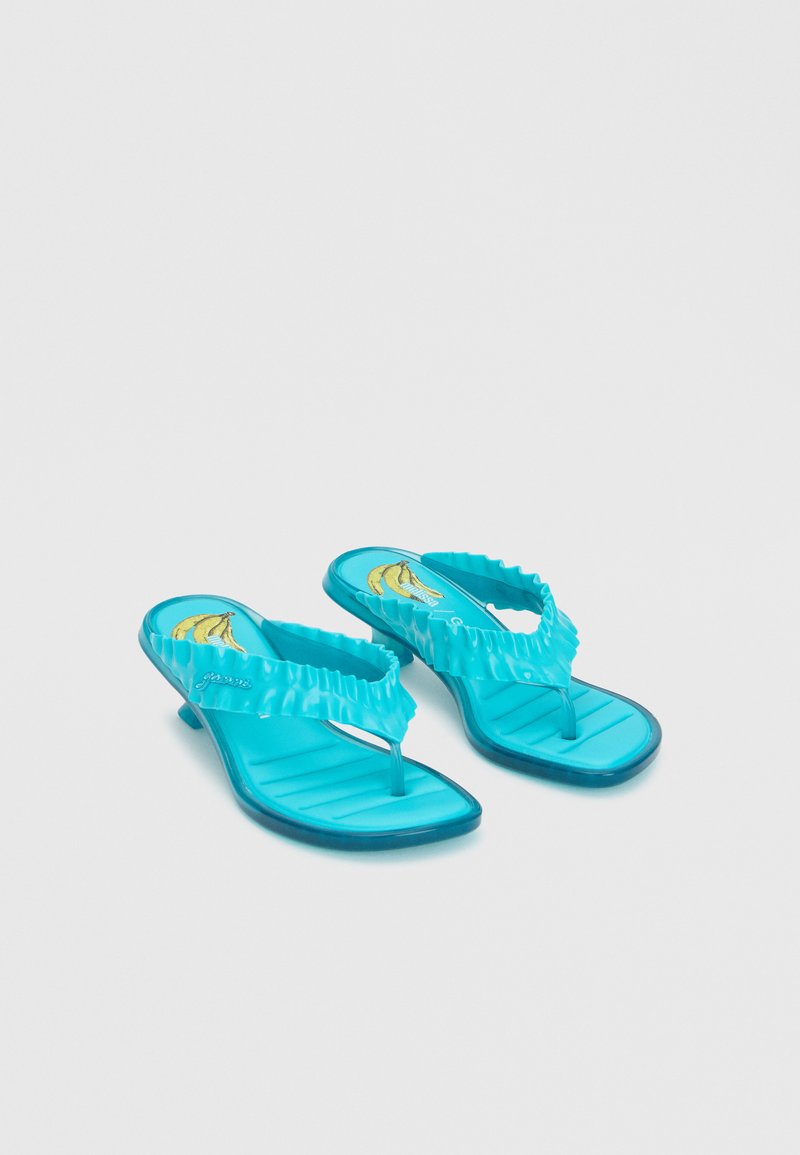 Paire de tongs à talons turquoise avec brides volantées et motif banane sur la semelle, présentée sur fond blanc.